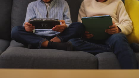 close up of two young boys sitting on sofa at home playing games or streaming onto digital tablet and handheld gaming device 1