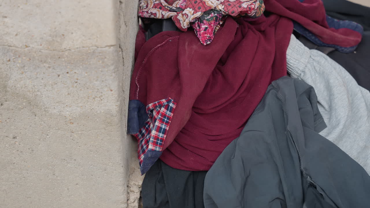 Clothes piled on concrete doorstep, soft sweaters and shirts left at rough stucco threshold, muted maroon and gray tones, closeup of fabric folds and frayed seams, empty entryway mood