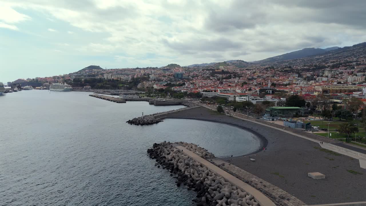 Slowmotion aerial drone footage of the city of Funchal with stone beach, curved breakwater, urban landscape and Atlantic coast on the island of Madeira, Portugal