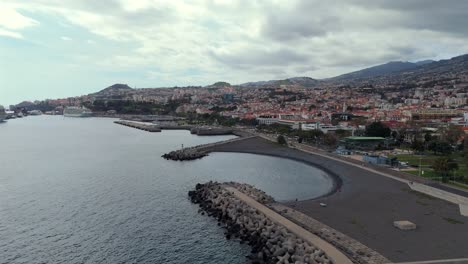 slowmotion aerial drone footage of the city of funchal with stone beach, curved breakwater, urban landscape and atlantic coast on the island of madeira, portugal