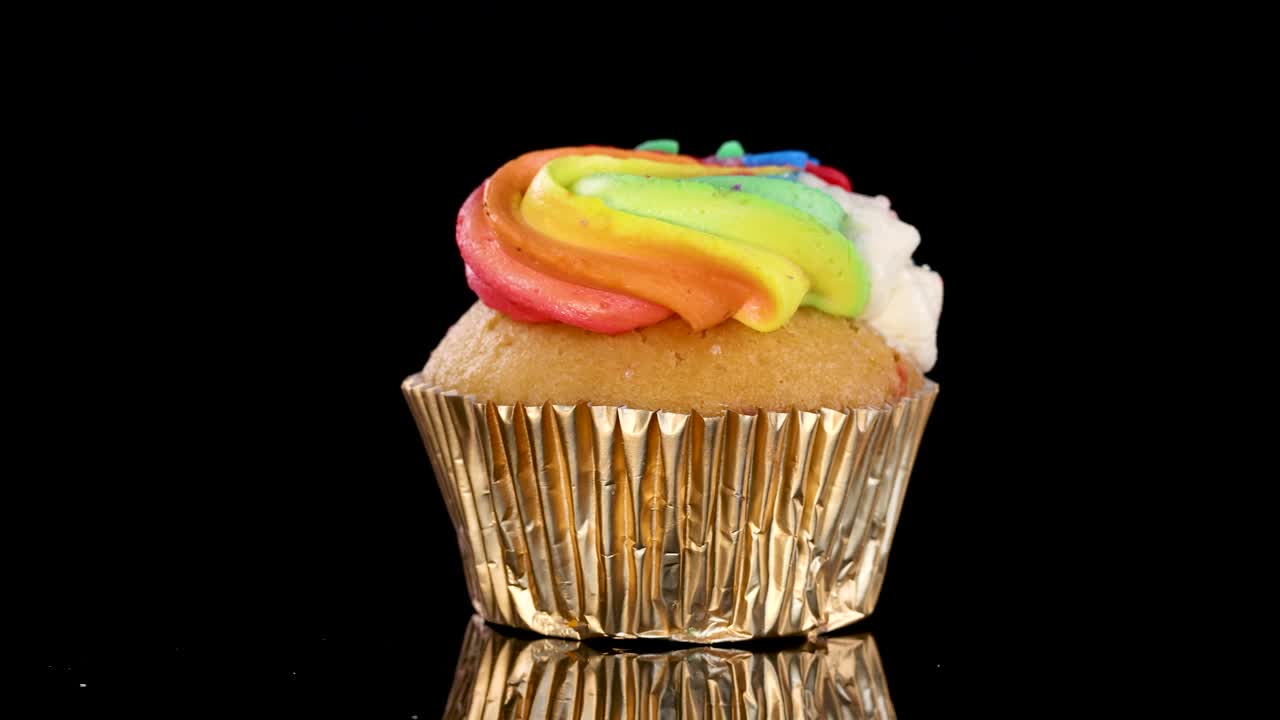 A single cupcake with rainbow frosting rotates smoothly on a reflective black surface, illuminated by studio lighting for a vibrant, appetizing presentation