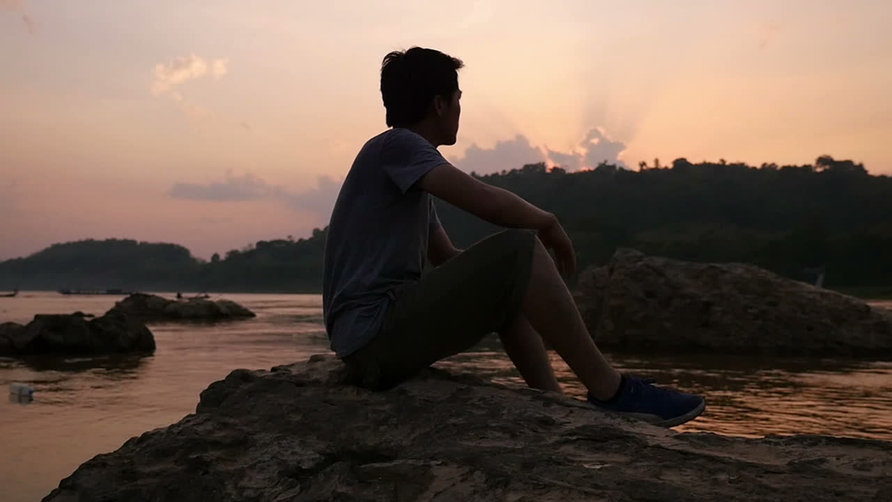 Uomo seduto su una roccia lungo un fiume al tramonto