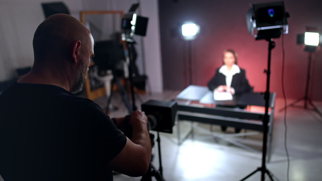 Mature man in black t-shirt stands at camera. Cameraman recording a video with female blogger.