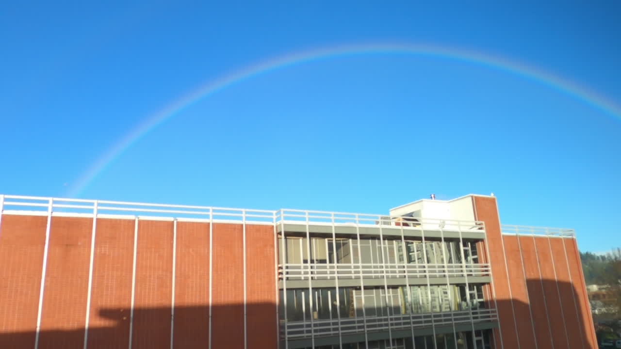A vibrant rainbow arches over a large brick building on a clear day