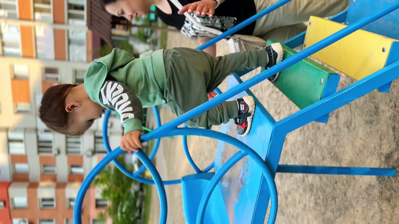 Cheerful Caucasian toddler going down from a slide. Mom secures her baby. Time spent at playground. Vertical screen.