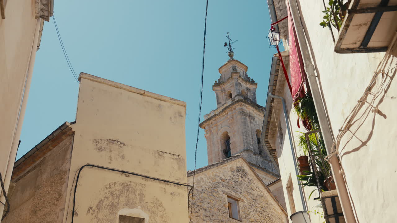 Una vista verso l'alto attraverso le strette vie di Bocairent, Spagna, rivela una storica torre campanaria incorniciata da case in pietra, balconi e la luminosa luce mediterranea.