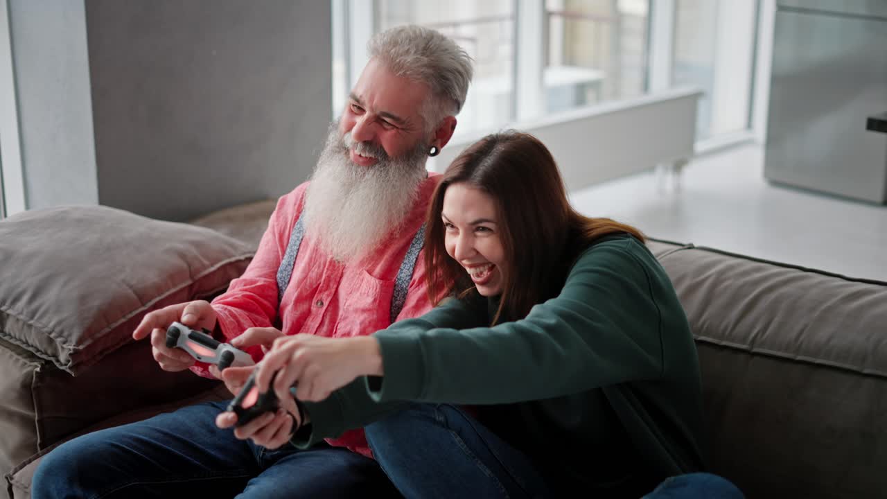 A brunette girl in a green sweater plays with her elderly father with gray hair and a lush beard in a video game using a joystick they have fun and rejoice while sitting on a modern sofa in an apartment with large windows overlooking the sea