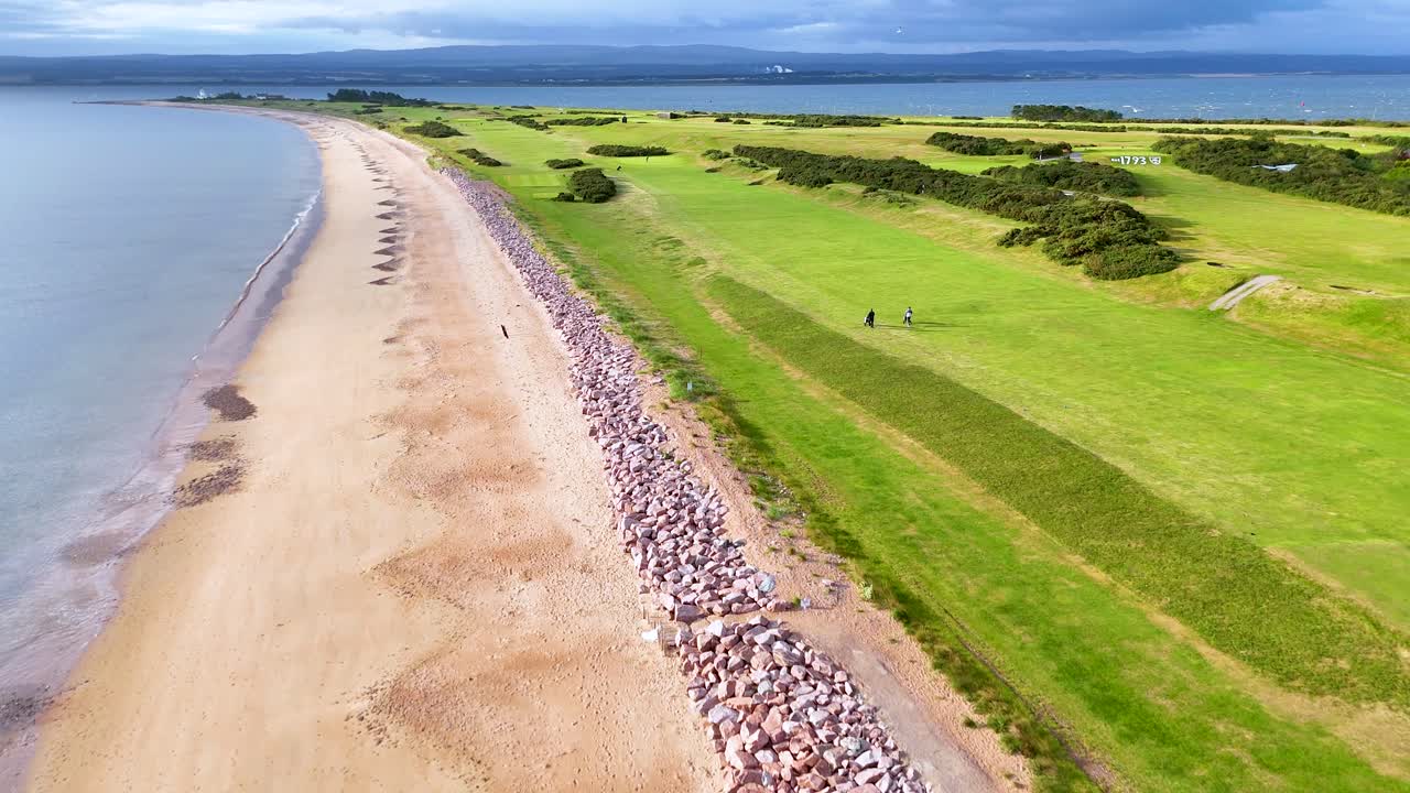 Drone footage glides above a sandy beach and adjacent golf course in Rosemarkie, Scotland, under bright daylight with sweeping, steady camera movement