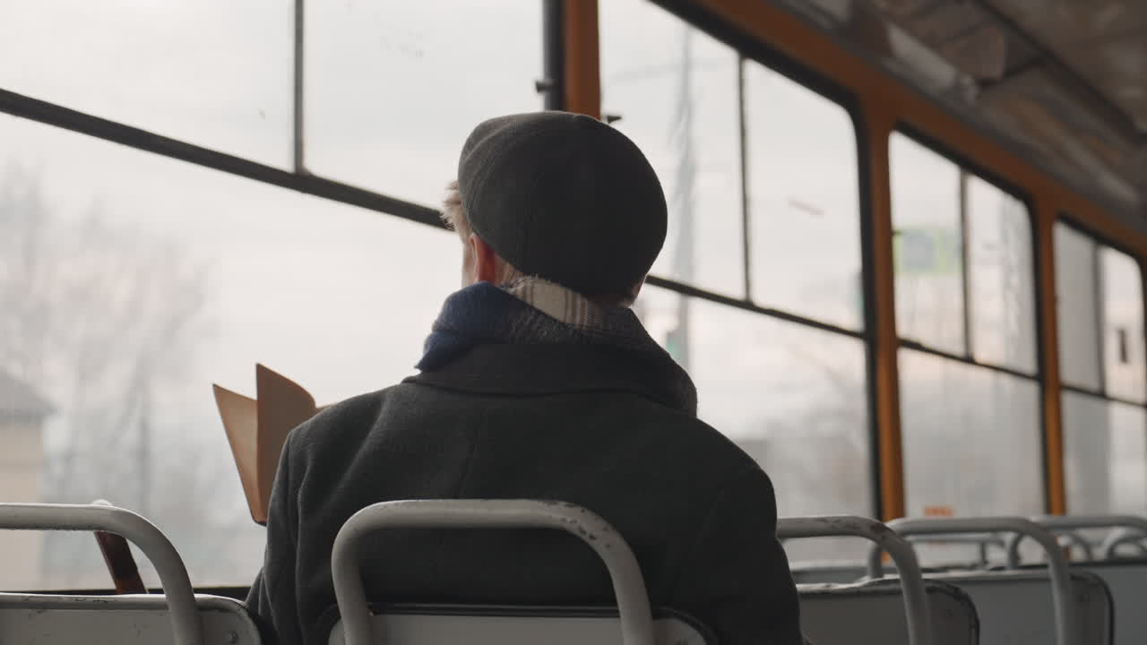 Creatore bianco su tram che studia un libro e si aggiusta il cappellino, pagine aperte contro un finestrino appannato. Giovane ricercatore bianco si gratta la testa, sfoglia le pagine e gesticola mentre legge, catturando un momento di quiete di riflessione su un problema.