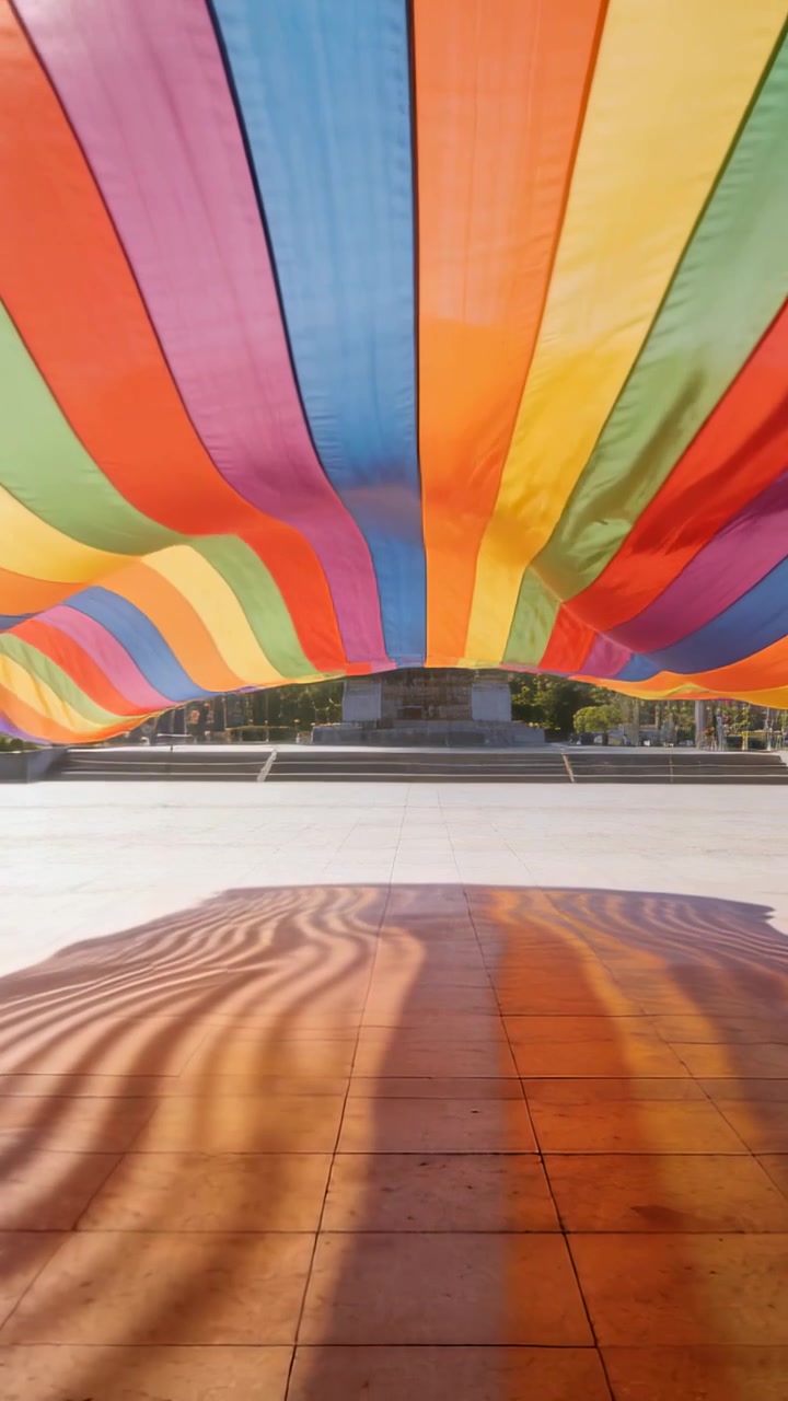 Vídeo vertical: Vento e sol levantando uma cobertura arco-íris que balança e projeta sombras vívidas em uma praça ladrilhada