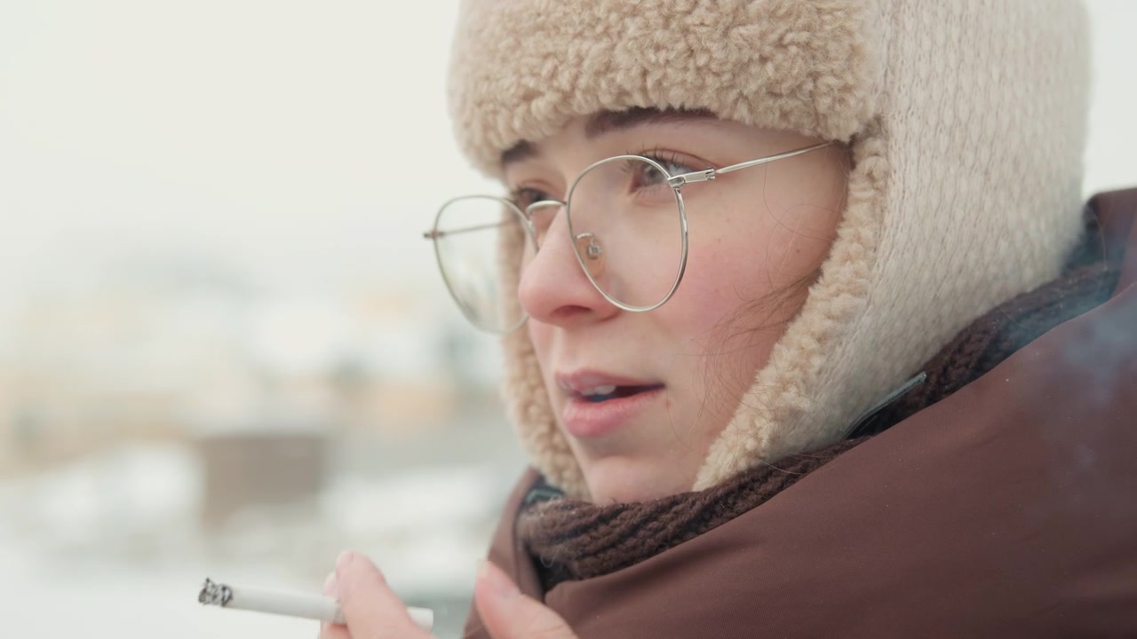 Primo piano di profilo a tre quarti di una giovane donna all'aperto in un ambiente urbano innevato, che tiene una sigaretta alle labbra con la mascella rilassata, respiro freddo visibile, cappello da trapper di lana, occhiali e luce diurna soffusa.