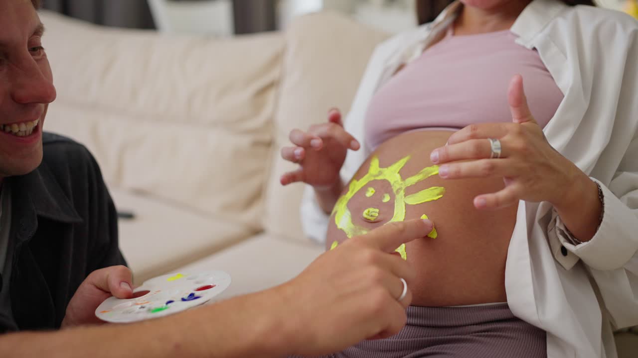 Close up of a happy pregnant woman drawing a sun on her belly while having fun with her husband at home on the sofa