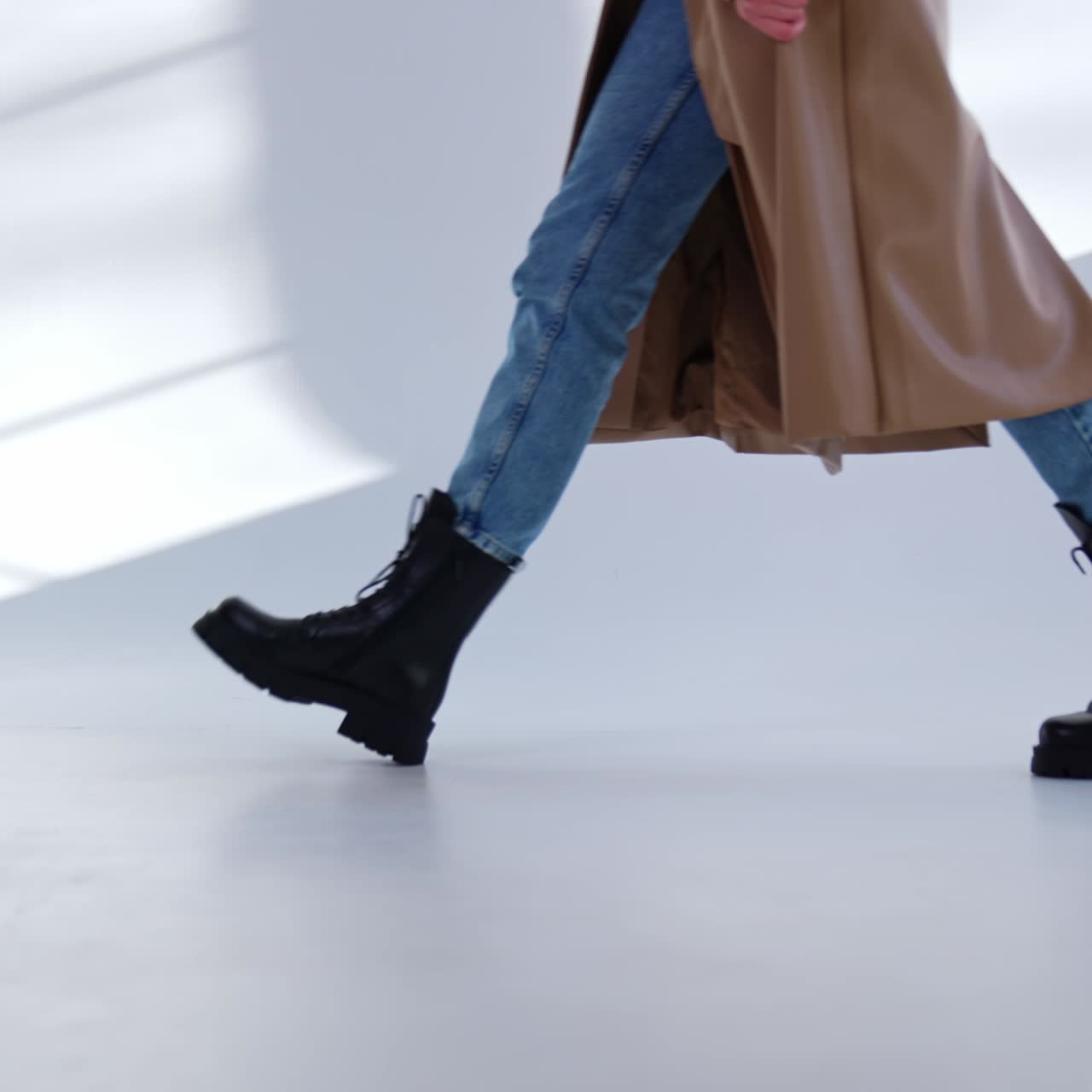 Unrecognized model wearing jeans, coat and boots walks by the white floor. Stylish black army boots demonstration