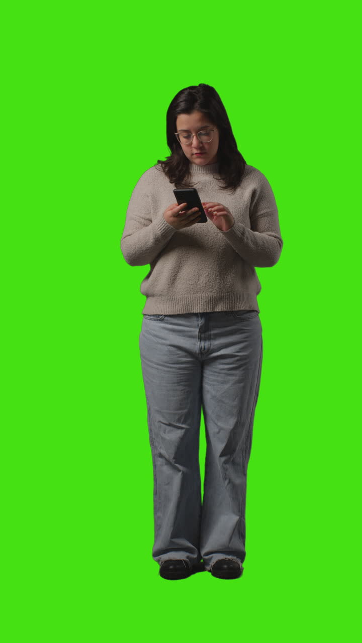 Video verticale: inquadratura a figura intera di giovane donna che usa il cellulare per messaggiare e navigare su internet, in piedi davanti a uno schermo verde.