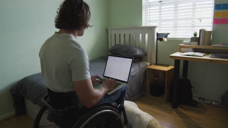 hombre caucásico discapacitado sentado en silla de ruedas usando una computadora portátil en el dormitorio