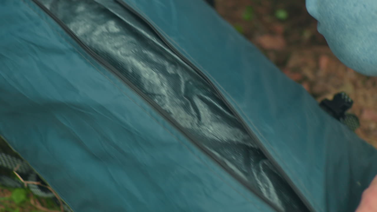 close up of person wearing sweater carefully unzipping green camping bag on forest ground surrounded by grass and leaves during outdoor trip, preparing for camping setup or packing session