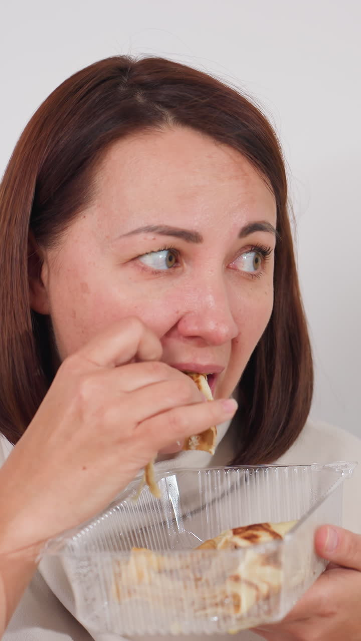 여자가 생각에 잠긴 표정으로 페이스트리를 맛보고 있어요. 차분한 스튜디오 부엌에서 투명한 플라스틱 용기에 담긴 페이스트리를 조심스럽게 한 입 베어 물고, 사려 깊은 시선으로 질감과 풍미를 음미하며 차분한 분위기를 연출합니다.