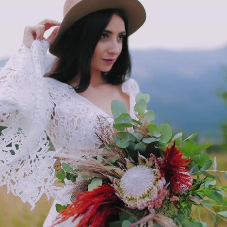 sexy girl with bouquet of flowers among nature. beautiful young woman in hat and white dress looking forward on blur mountains background.