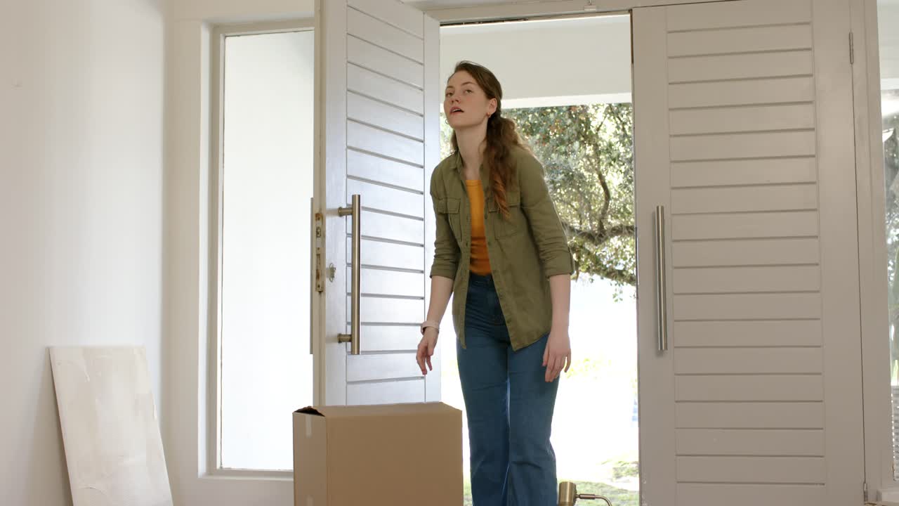 Woman carrying cardboard box stepping inside foyer with doors opening, surveying new home