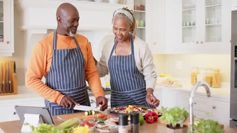african american senior couple in aprons preparing meal using tablet in kitchen, slow motion