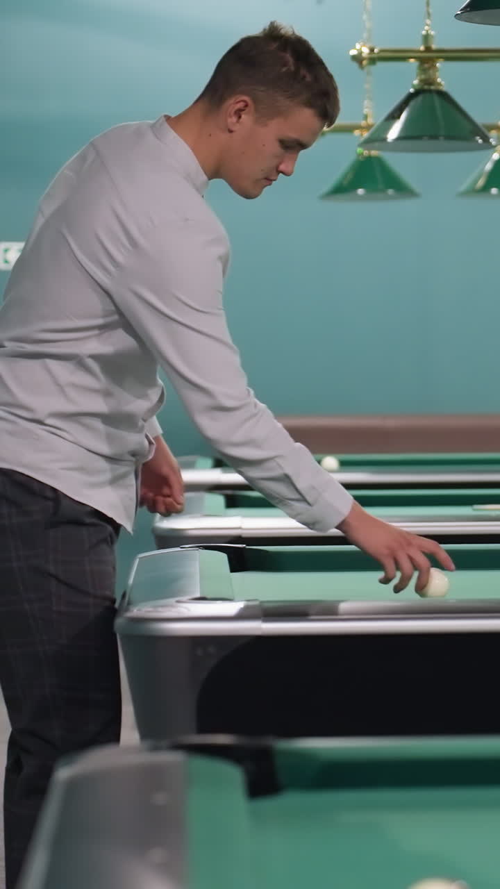 joven con camisa blanca y pantalones grises entrando en una sala de billar con mesas de billar verdes y lámparas superiores expresión enfocada, entorno moderno de club deportivo, atmósfera de ocio elegante