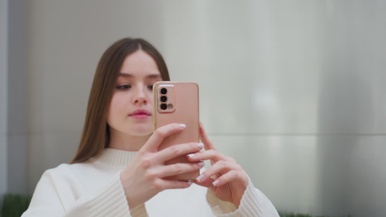 Lady in white sweater taking picture with smartphone, focused expression, modern interior background with soft lighting and elegant design, minimalist setting with green decorative plants