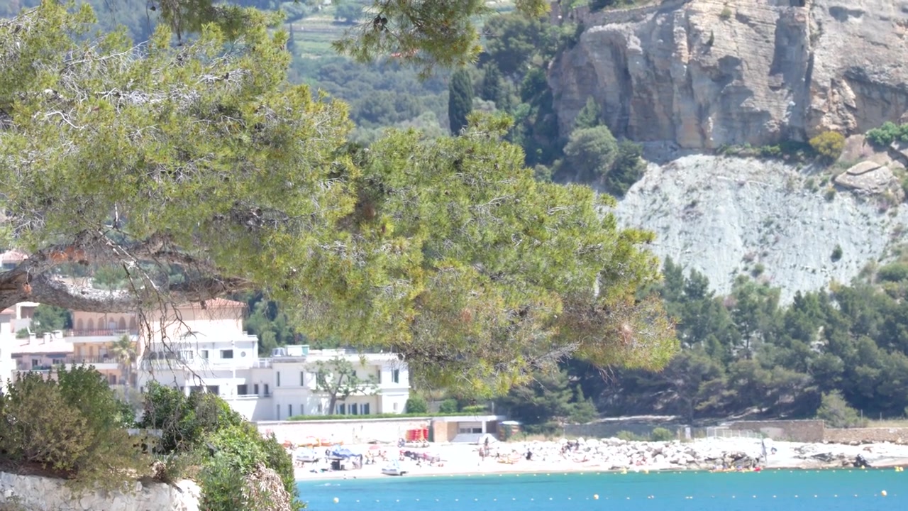 Static wide shot of a coastal village nestled against limestone cliffs under bright daylight.