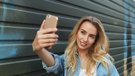 joven mujer rubia caucásica con chaqueta de jeans tomando un selfie en el teléfono inteligente en la calle