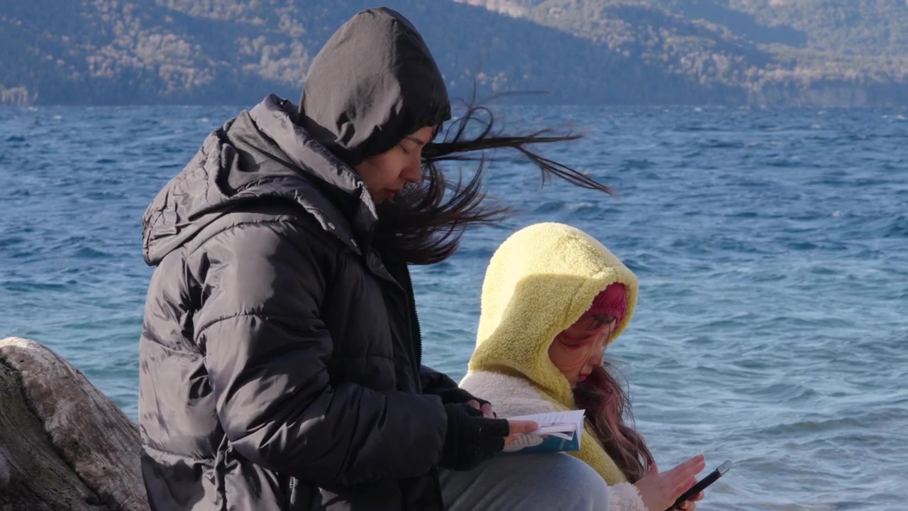 A mother and daughter engrossed in reading by a cold lakeside. Ideal for depicting family moments or outdoor leisure activities in winter settings.