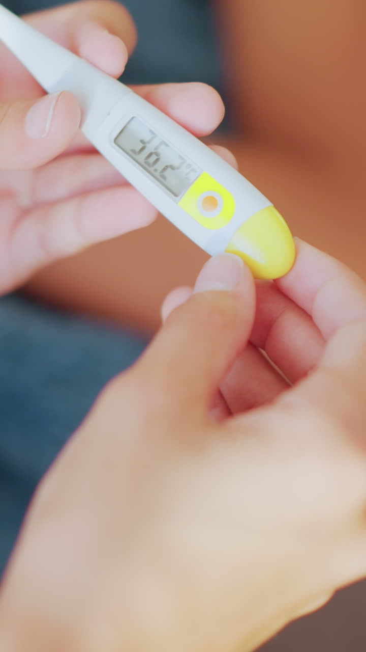 Close up of hands holding thermometer with clear digital display reading 36 degrees Celsius while someone lies down blurred in background showing monitoring care and health check