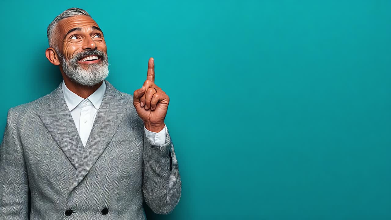 En glad mand med sølvfarvet hår og skæg i et gråt jakkesæt, der peger opad mod en lys turkis baggrund, udstrålende positiv energi og inspiration.