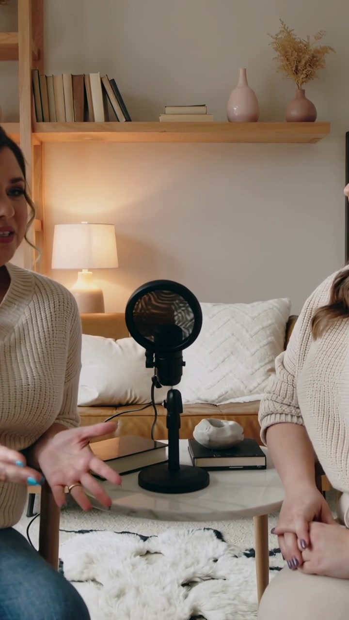 Vertical video: Speaking left woman wearing knits gesturing to mic on table at home for podcast