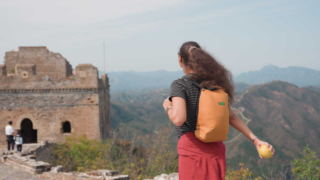 Zainatrice che ammira il panorama montano, zaino giallo e cappello, in piedi vicino all'ingresso di una torre di guardia, muro serpeggiante che si perde in lontananza, brezza stagionale, giacca rossa legata in vita, postura rilassata, viaggio
