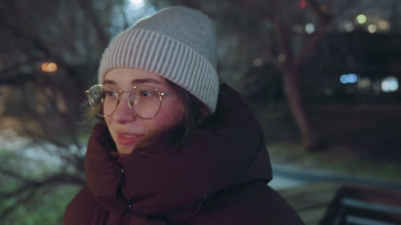 White woman thoughtful smile under lights in park, round glasses catching lamplight, knitted hat and puffer jacket, subtle expression changes from shy to playful, cinematic grain and soft nighttime