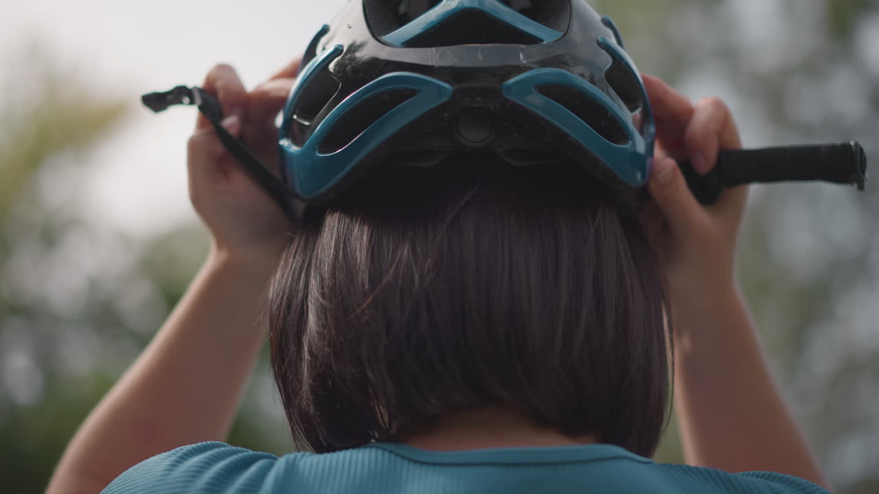 Donna che regola il casco da bicicletta all'aperto, profilo posteriore che mostra un taglio di capelli a caschetto e una maglia verde acqua, allacciando il cinturino e la visiera, cielo nuvoloso con alberi sfocati, stato d'animo calmo e concentrato, pendolare che si prepara per l'uso individuale