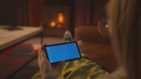 Mujer-En-Casa-Sentada-En-Un-Sofá-Con-Fuego-Transmitiendo-A-Un-Teléfono-Móvil-De-Pantalla-Azul-Sosteniendo-Una-Copa-De-Vino