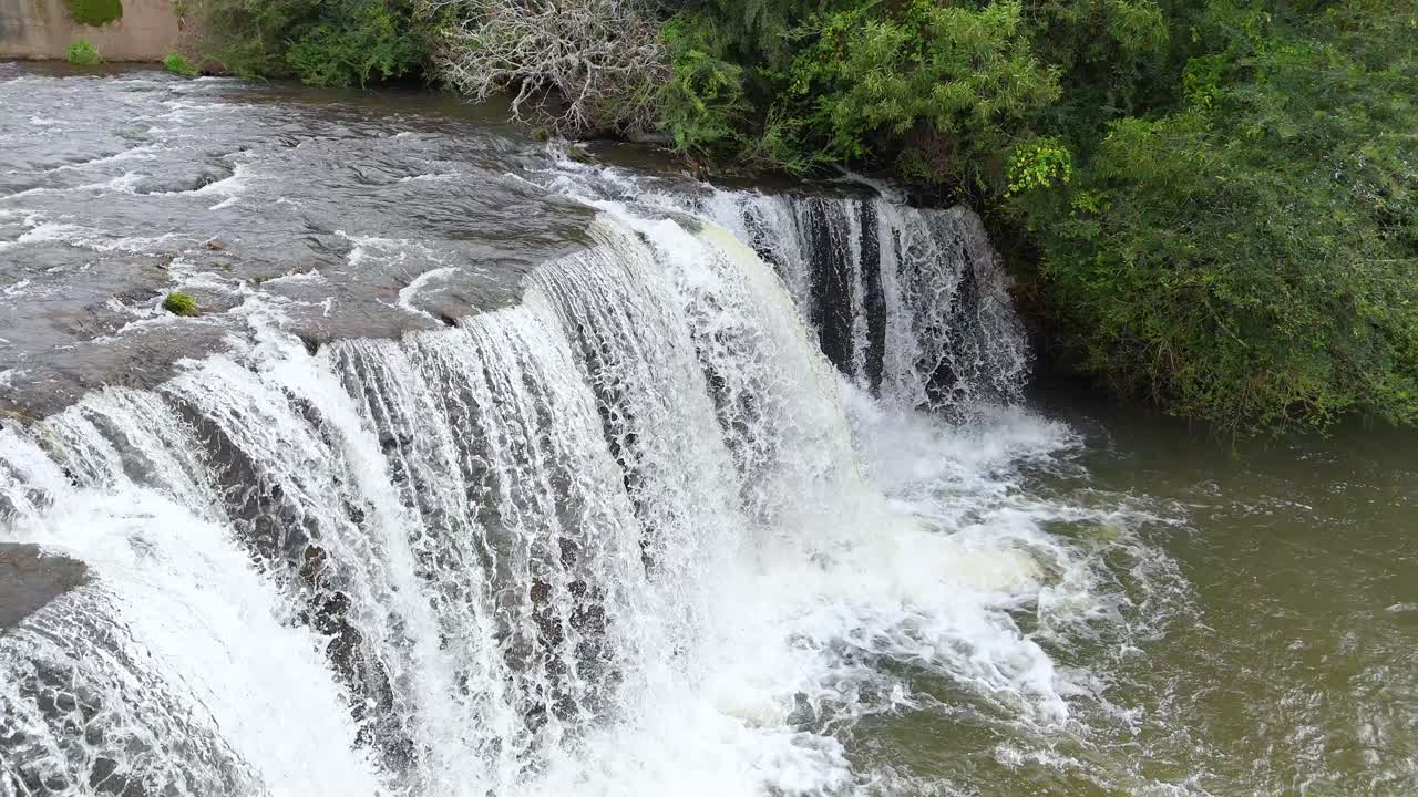 Drone captures steady overhead shot of wide waterfall, lush greenery, and flowing river in daylight