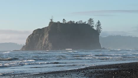 aerial drone footage of a tree-covered sea stack rising from the ocean along a rugged coastal beach