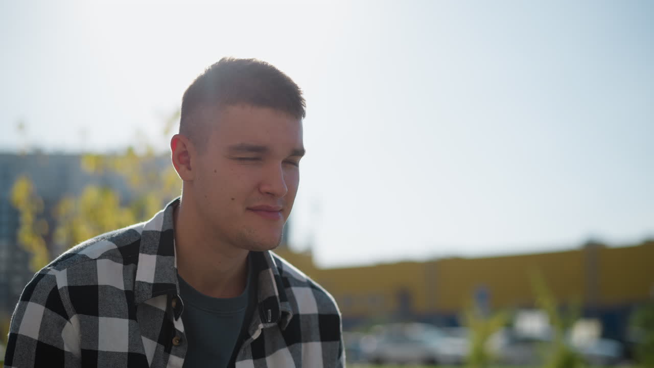 fair skinned man in checkered shirt puts sweet in mouth while enjoying calm sunny day outdoors with soft blur background featuring parked cars, trees, and yellow building in afternoon light