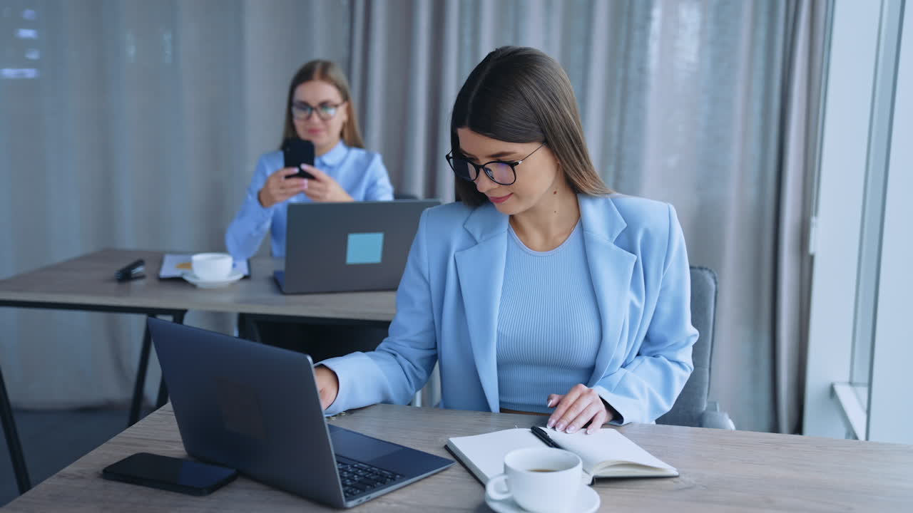 Young female ladies sitting at desks in front of laptops. Employees using gadgets in modern office. Blurred backdrop.