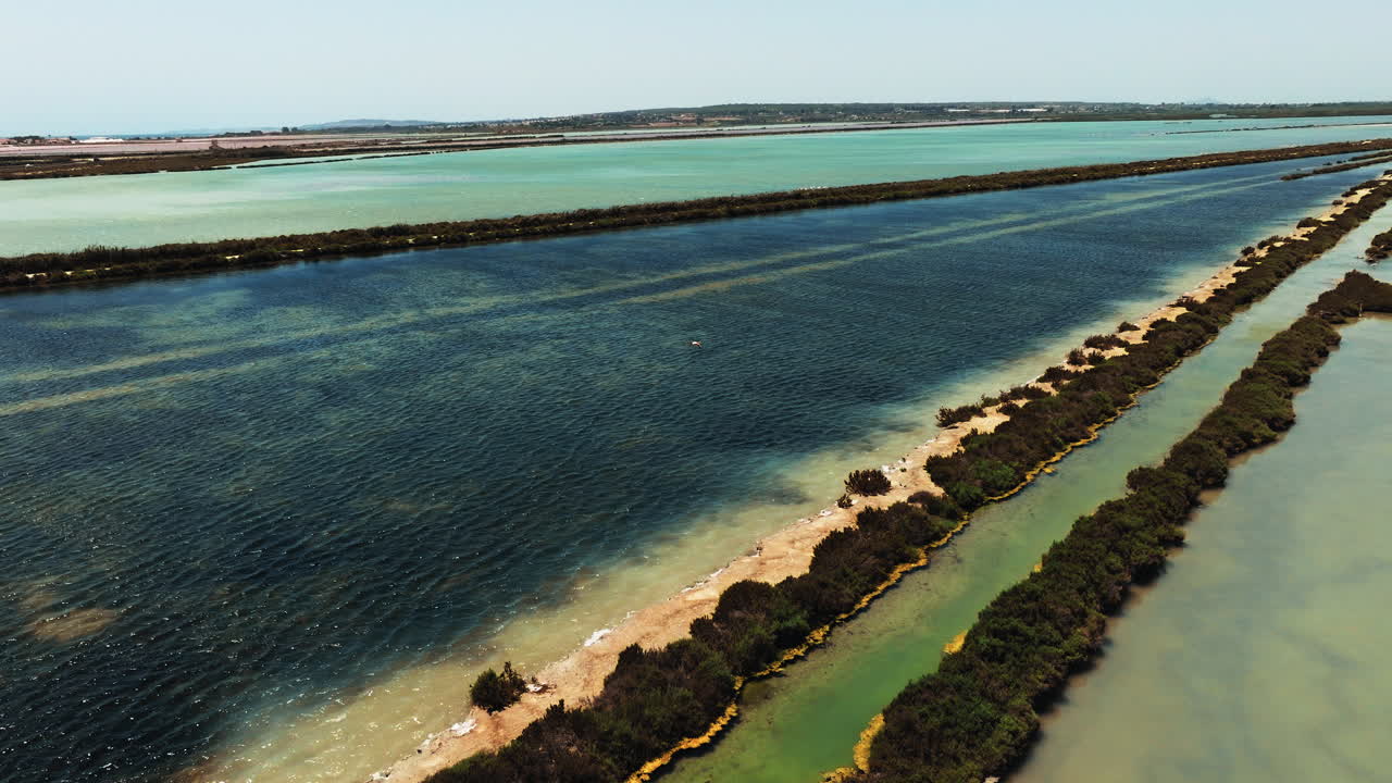 Drone flyover of the Santa Pola salt fields near Alicante, Spain, showing canals and vibrant blue and green salt lakes across the Mediterranean landscape