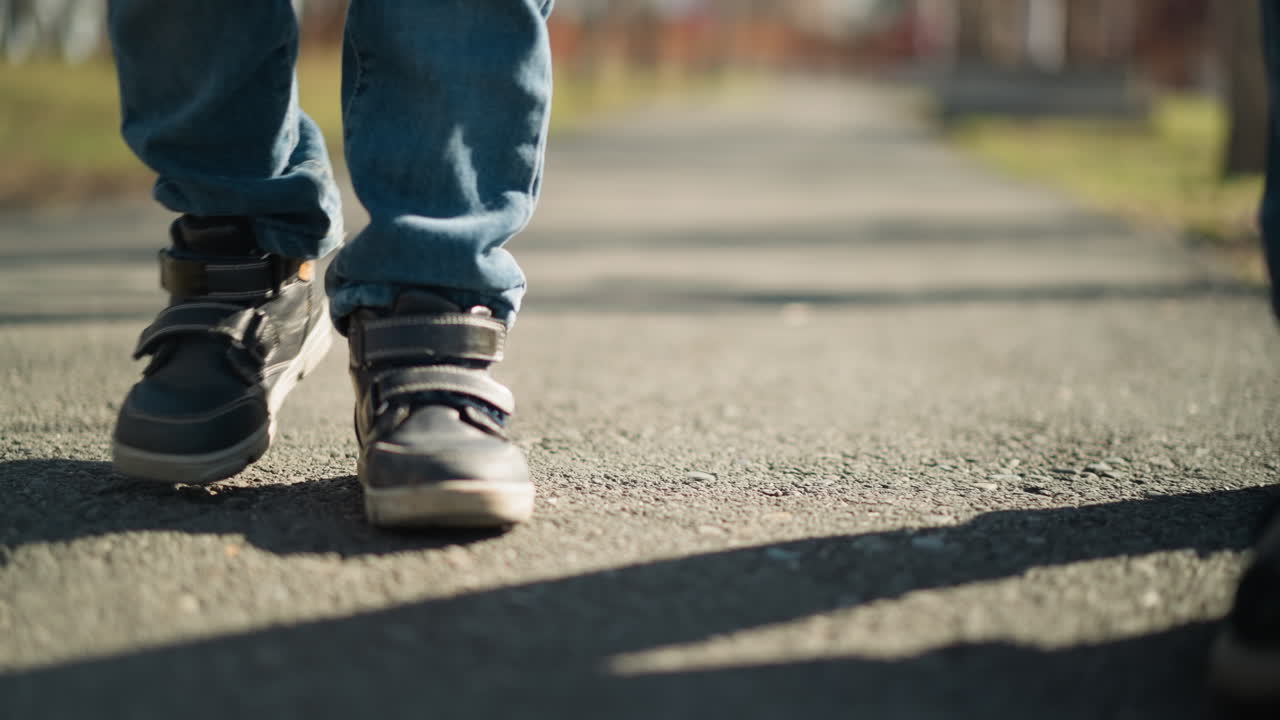un primer plano de un niño y un adulto con vaqueros y botas caminando por una acera, la cámara captura sus sombras reflejadas en el suelo mientras caminan