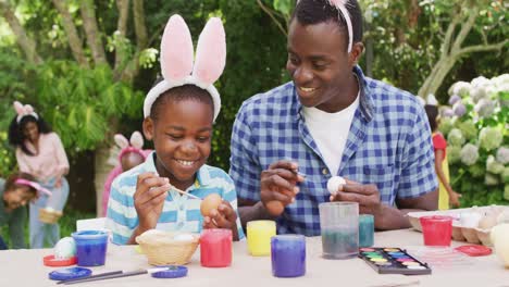 animation of happy african american father and son painting easter eggs in garden