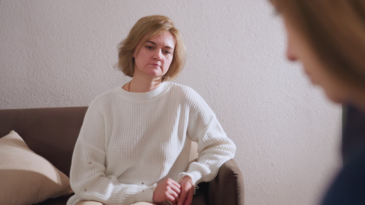 Mindful guest in white sweater seated attentively on sofa listening with calm focus during support session, showing emotion and engagement in conversation with specialist in soft lit room