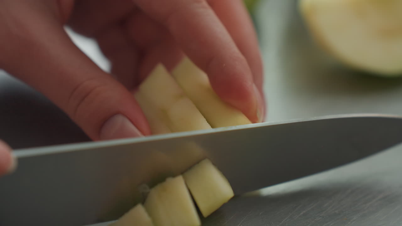 Manos cortan pera en dados. Cortando cuidadosamente pera en superficie de acero inoxidable. Cortando pera con precisión en tabla de cortar limpia en la cocina. Ambiente de cocina tranquilo con manos preparando hábilmente fruta para ensalada