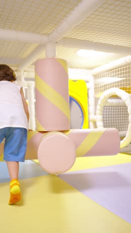blond young girl actively engaged in an indoor playground, intently spinning a large, soft marshmallow-shaped obstacle with pink and yellow stripes