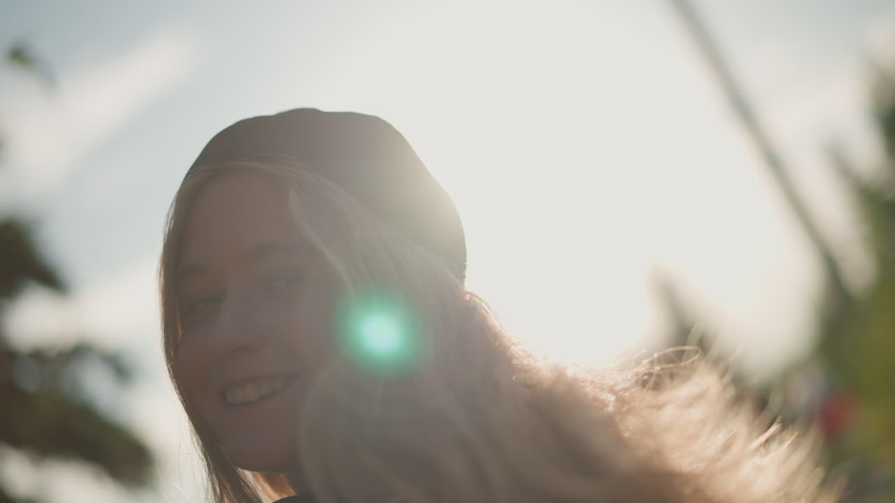Retrato com fones de ouvido em rua urbana ensolarada com luz de fundo dourada suave. Mulher jovem usando boné e fones de ouvido no pescoço, alcançando a câmera de forma divertida, bokeh quente, sorriso relaxado, vibe de música indie
