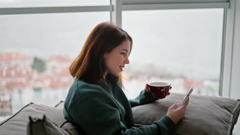 Happy-brunette-girl-on-a-green-sweater-sits-on-a-modern-sofa-drinks-tea-from-a-brown-mug-and-scrolls-through-her-social-media-feed-in-a-White-phone.-Sitting-in-a-modern-apartment-with-large-windows-overlooking-the-sea