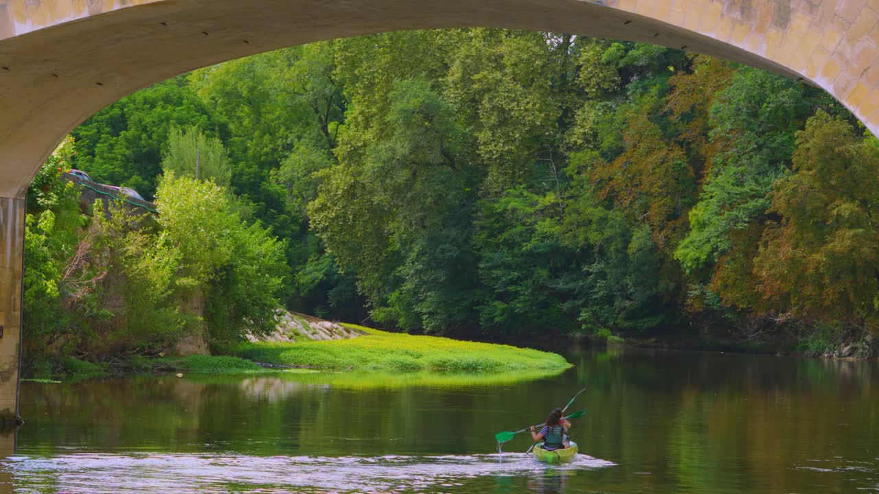 kayakers turísticos irreconocibles remando en cámara lenta en el tranquilo río vézère bajo el puente medieval histórico en montignac, francia, en un caluroso día de verano con árboles verdes exuberantes.
