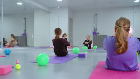 sesión de entrenamiento en grupo en un gimnasio bien iluminado con los participantes rodando hacia atrás en esteras, manos juntas en las rodillas, bajo el ojo atento de un instructor reflejado en un gran espejo de gimnasio